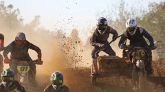 Sidecarcross racing action with professional riders navigating a dirt track, featuring intense roost and mud spray under bright sunlight