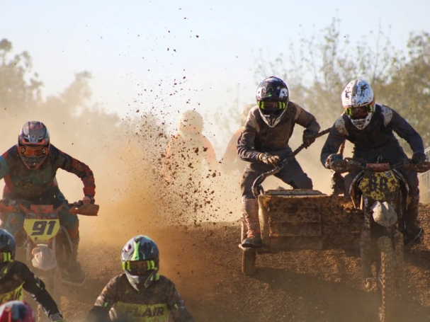 Sidecarcross racing action with professional riders navigating a dirt track, featuring intense roost and mud spray under bright sunlight