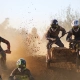 Sidecarcross racing action with professional riders navigating a dirt track, featuring intense roost and mud spray under bright sunlight