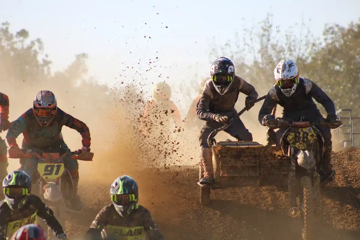Sidecarcross racing action with professional riders navigating a dirt track, featuring intense roost and mud spray under bright sunlight