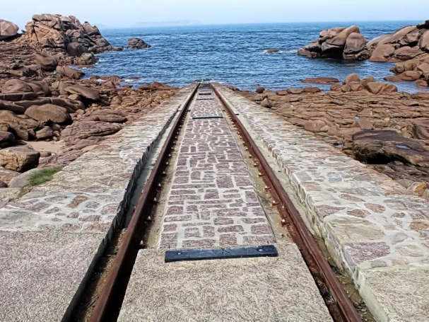 Lifeboat launch ramp with rusty rails leading into the sea, surrounded by pink granite rocks in Ploumanac'h, Brittany.