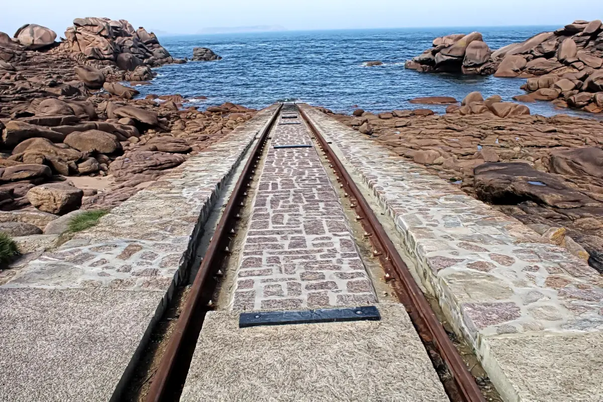 Lifeboat launch ramp with rusty rails leading into the sea, surrounded by pink granite rocks in Ploumanac'h, Brittany.