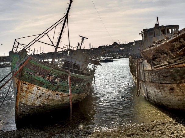 Two weathered wooden shipwrecks at the Camaret-sur-Mer boat graveyard, resting on a pebble beach at low tide under a hazy sky