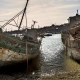 Two weathered wooden shipwrecks at the Camaret-sur-Mer boat graveyard, resting on a pebble beach at low tide under a hazy sky