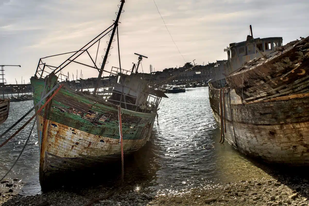 Two weathered wooden shipwrecks at the Camaret-sur-Mer boat graveyard, resting on a pebble beach at low tide under a hazy sky
