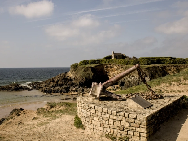A large rusted memorial anchor at Le Pouldu, Brittany, with the historic customs house on the cliff overlooking the Atlantic coast