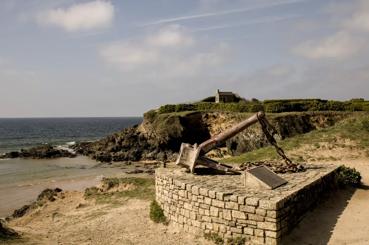 A large rusted memorial anchor at Le Pouldu, Brittany, with the historic customs house on the cliff overlooking the Atlantic coast