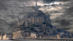 Wide view of Mont-Saint-Michel abbey under a dramatic, cloudy grey sky with a wooden footbridge in the foreground