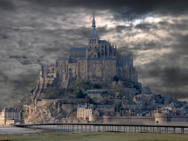 Wide view of Mont-Saint-Michel abbey under a dramatic, cloudy grey sky with a wooden footbridge in the foreground