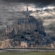Wide view of Mont-Saint-Michel abbey under a dramatic, cloudy grey sky with a wooden footbridge in the foreground