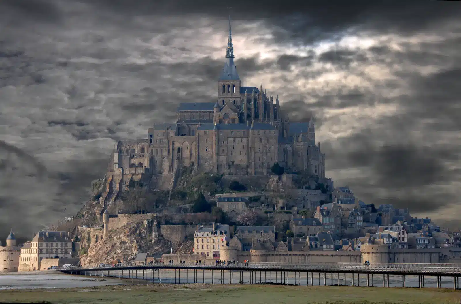 Wide view of Mont-Saint-Michel abbey under a dramatic, cloudy grey sky with a wooden footbridge in the foreground