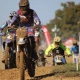 Close-up of a muddy motocross rider leading a pack of competitors on a dirt track."