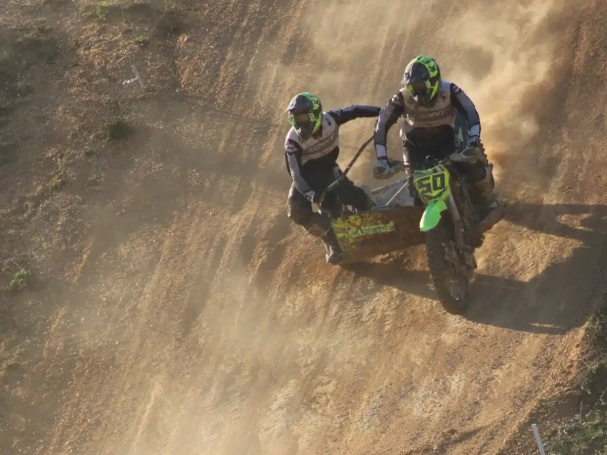 Sidecarcross team leaning hard during a steep downhill descent on a sandy track with backlight dust
