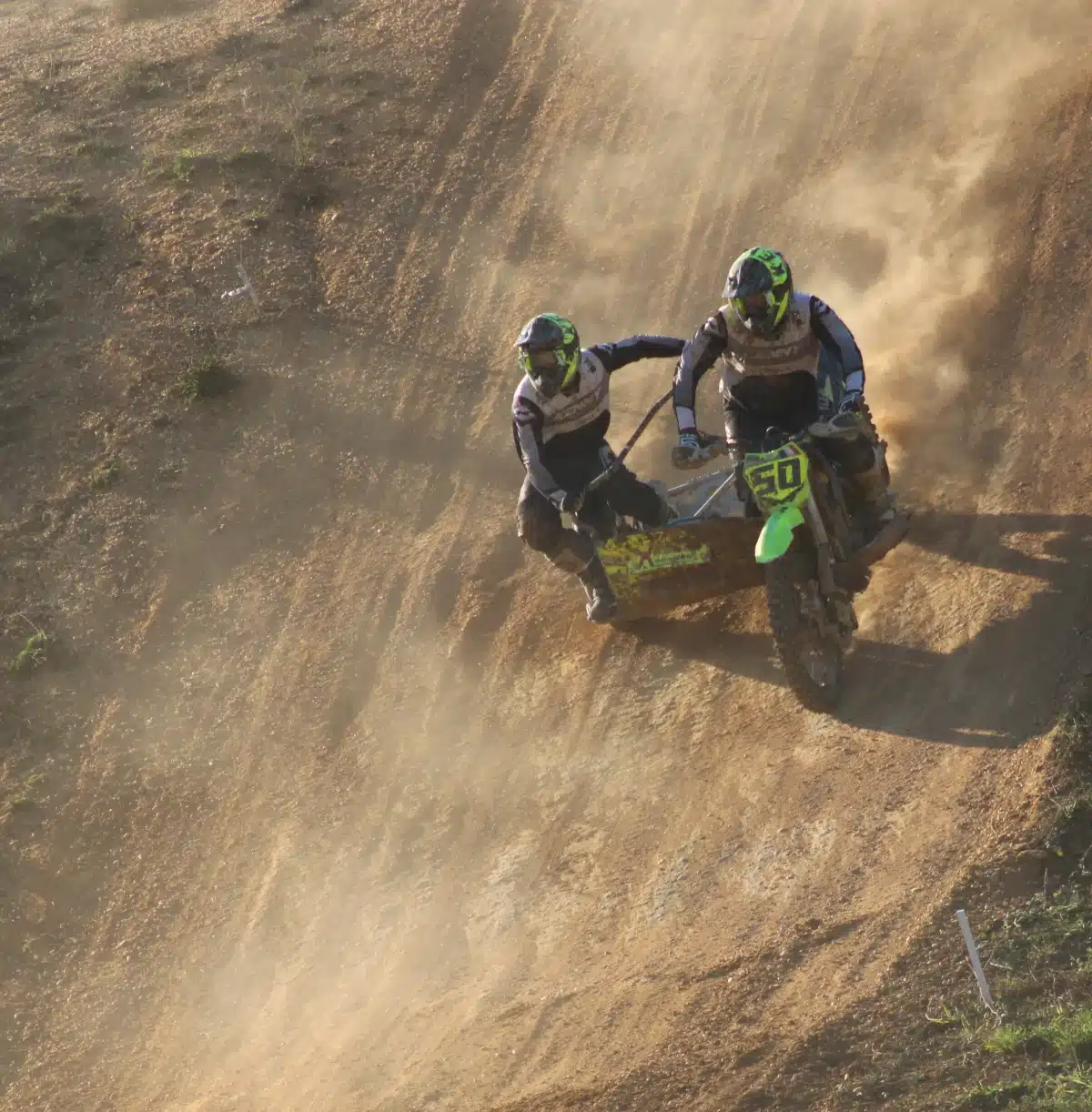 Sidecarcross team leaning hard during a steep downhill descent on a sandy track with backlight dust